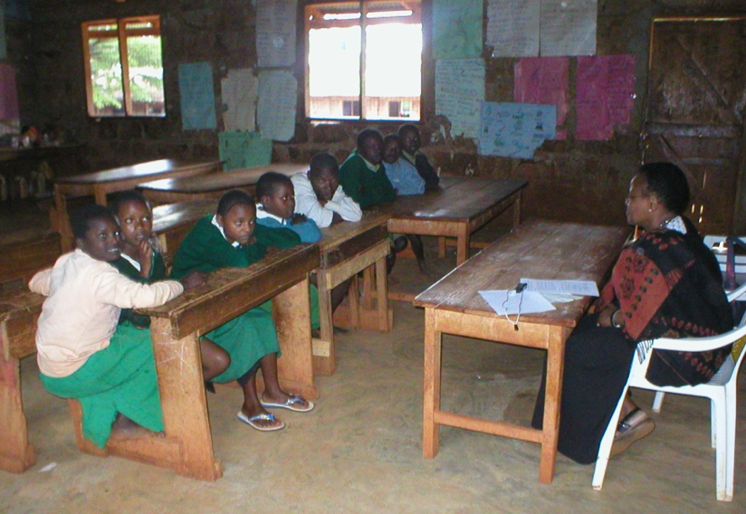 A number of students in a classroom sitting at their desks and a teacher at a teacher desk in front